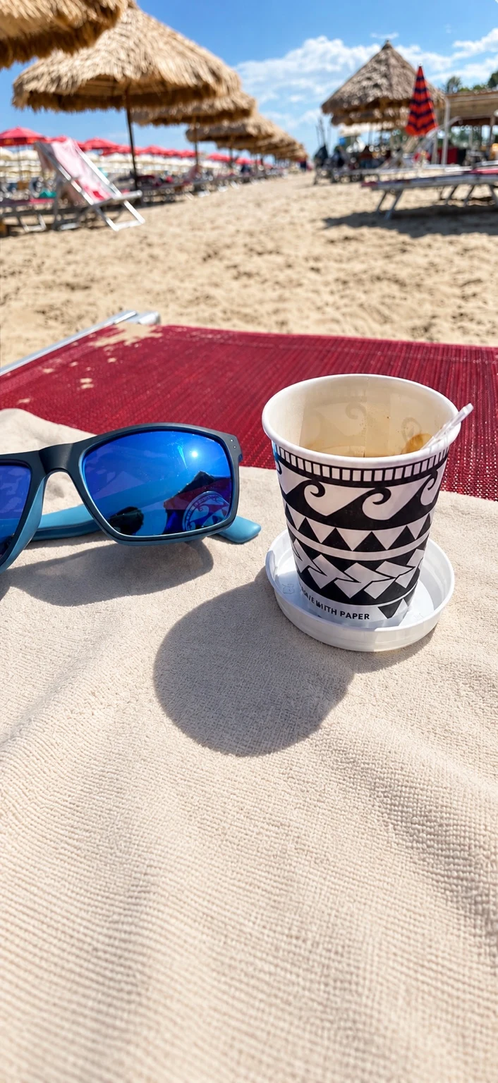 A coffee cup and blue sunglasses on a light beach towel, with sandy beach and straw parasols beyond